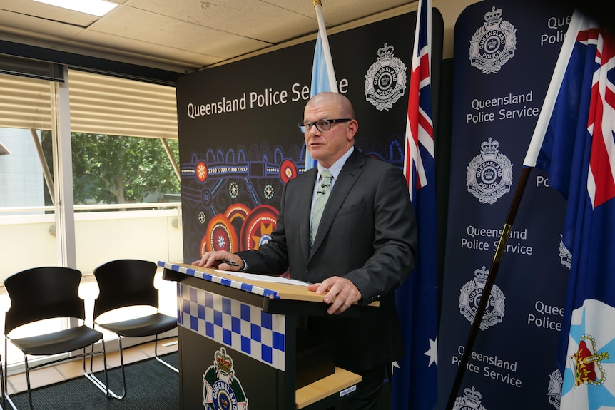 A man in a suit and tie, standing behind a podium with Queensland police parephenalia. 