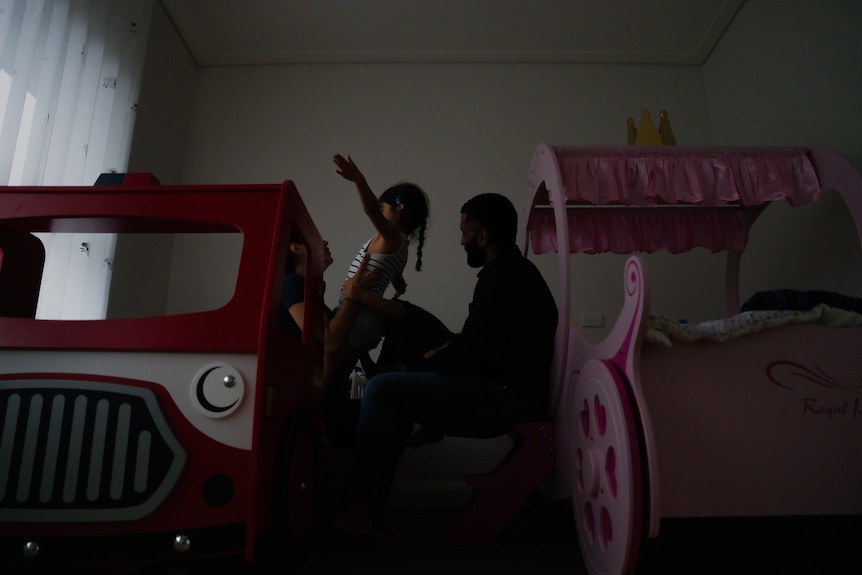 A mother, father and two children huddle together between two colourful beds. One child leans back, arms outstretched.