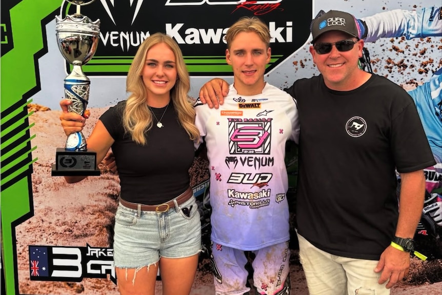 A young lady stands next to a young man in motocross gear who is also next to their father.