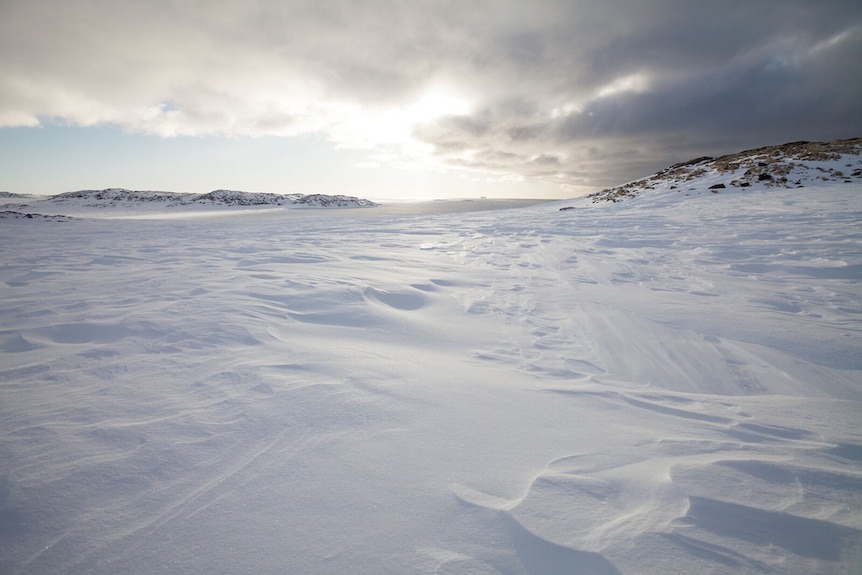A landscape shot dominated by a vast icy expanse, with grey clouds and a glimmer of sunlight above