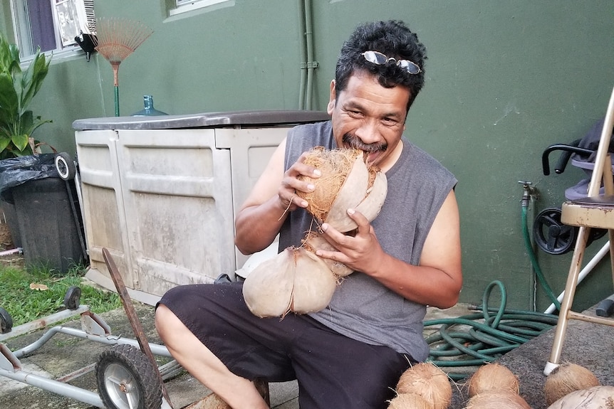 Man from marshall islands wearing grey tank top, sunglasses on head, and biting into coconut husk