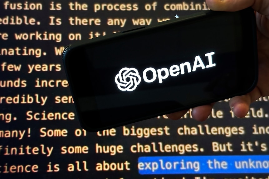 The OpenAI logo on a mobile phone in front of a screen showing part of the company website.