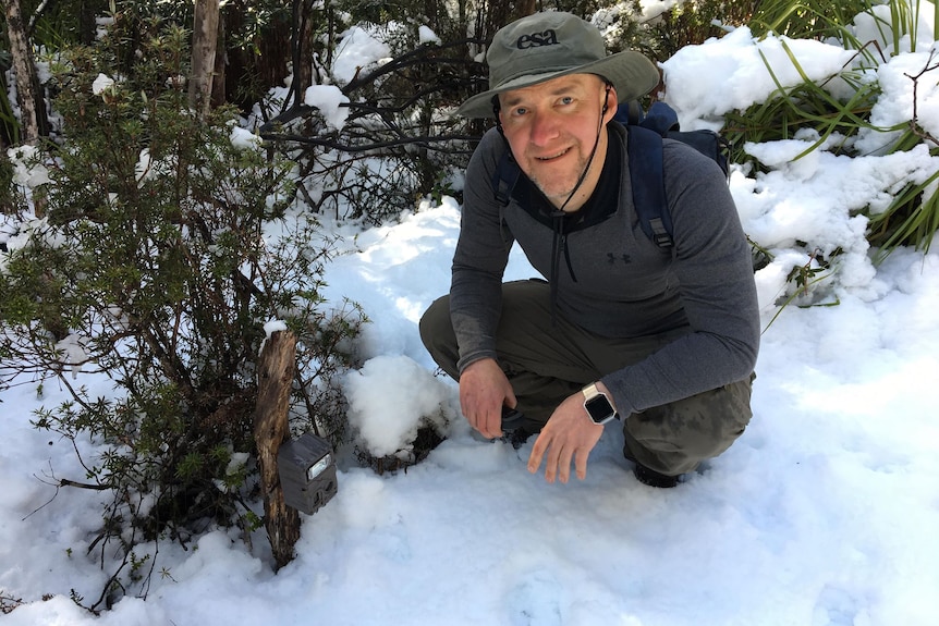 A man crouches down in the snow next to a small camera mounted on a wooden pole