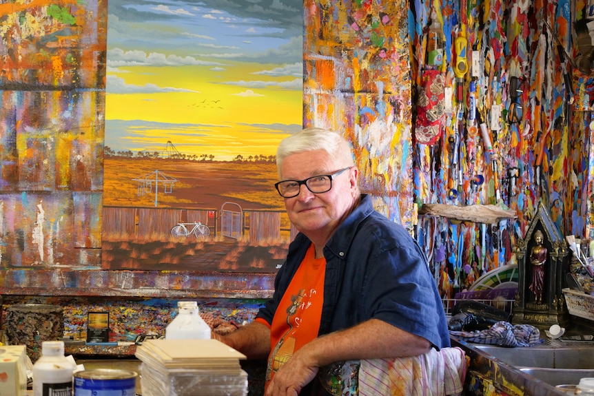 An older man in a shirt and glasses sitting in front of a painting of a horizon and a wooden wall splattered with colours
