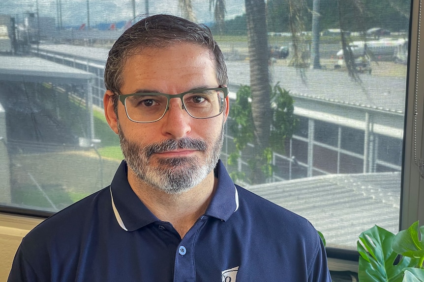 Man with beard, glasses and blue polo in office with airport in background