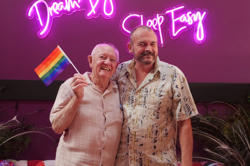 Two men smile with one holding a rainbow flag.