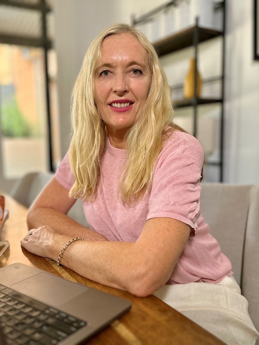 woman with long blonde hair and pink tshirt smiling at desk with computer