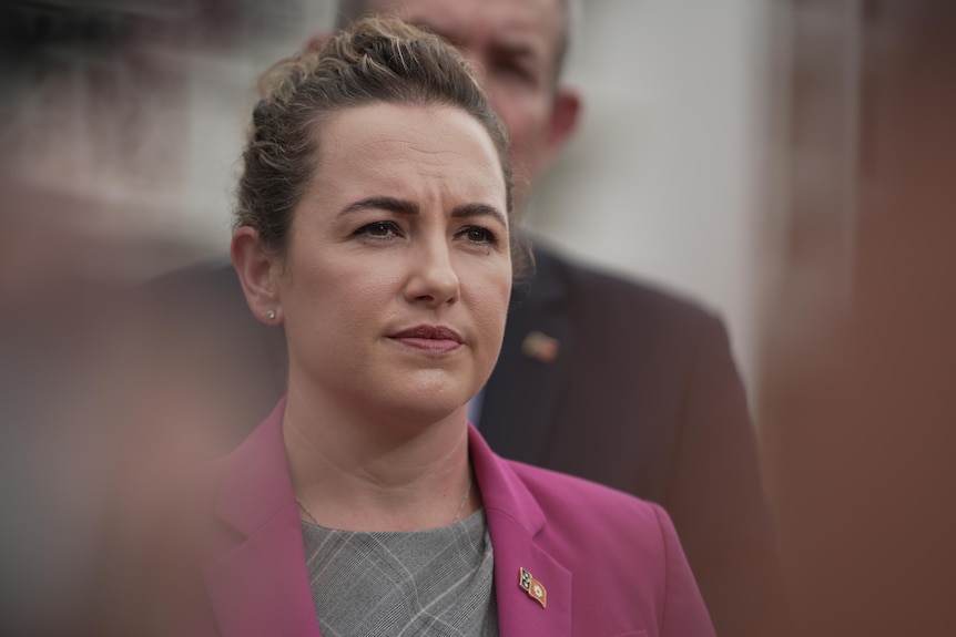 A woman in a pink blazer stares into the middle distance.