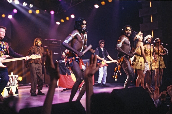 Nine people on stage, some of whom are wearing traditional First Nations body paint and dress, singing into microphones.