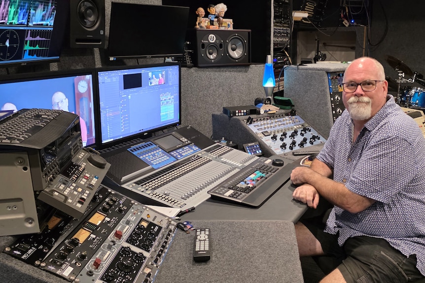 Man with a white beard and glasses sitting in a studio in front of buttons and screens.