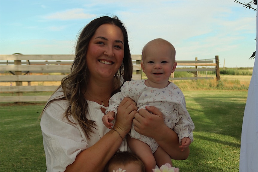 A smiling woman with long hair holds a smiling baby while standing in a yard on a farm.