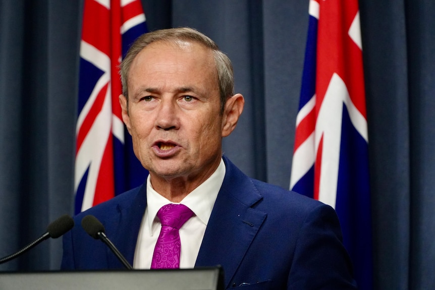 A close-up shot of WA Premier Roger Cook speaking at a media conference indoors, wearing a suit and tie, flags behind him..