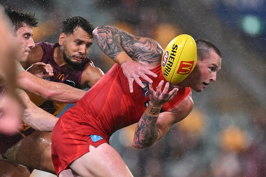 A Gold Coast Suns player leans forward as he tries to keep hold of the ball while being chased by two defenders.