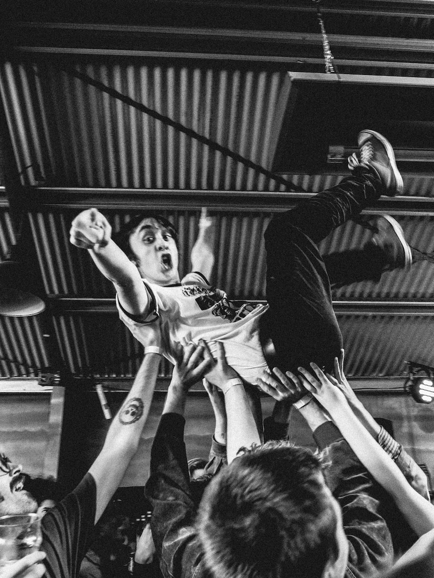 Black and white photo of a chaotic music event with a young man crowdsurfing across a lively crowd