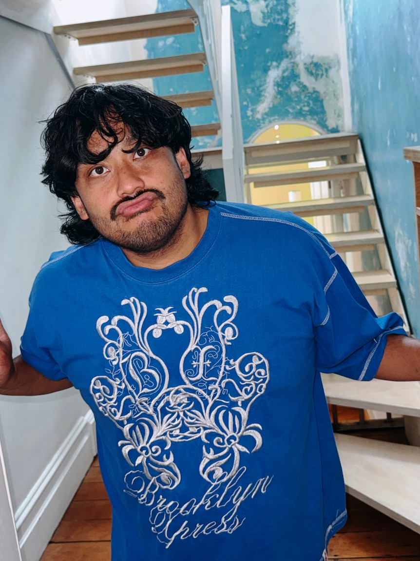 David Correos, a Filipino Kiwi with a moustache in a royal blue T-shirt, stands in front of a staircase, pouting.