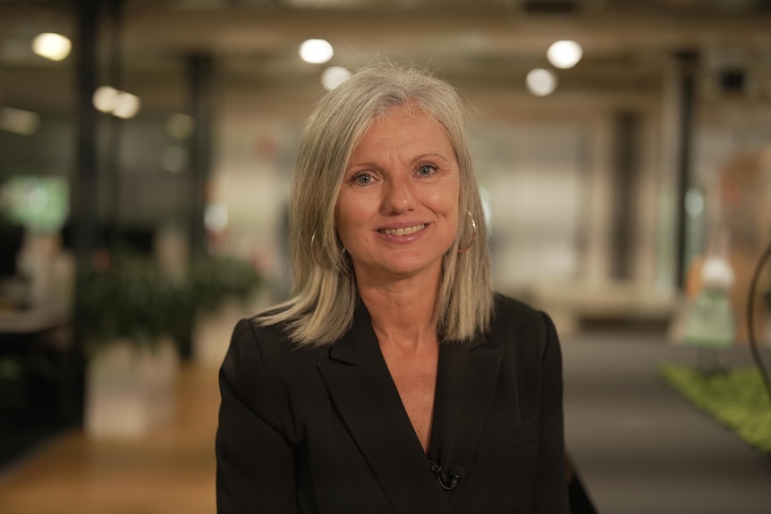 A woman in an office in a black suit smiles at the camera.
