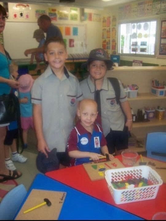 Three children stand together in uniforms in a classroom smiling.