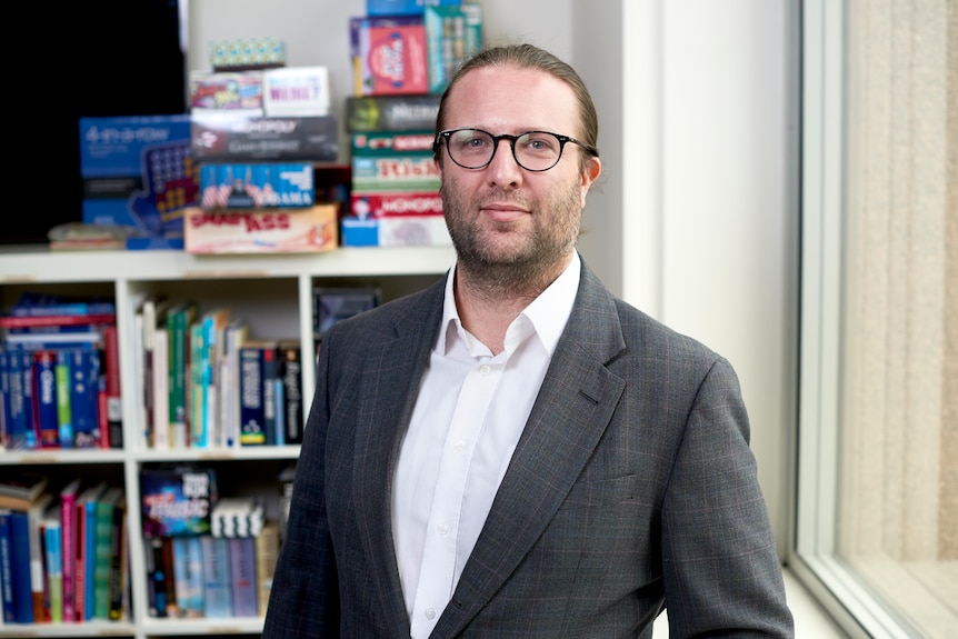 Tom Sulston stands in front of a book shelf and smiles at the camara