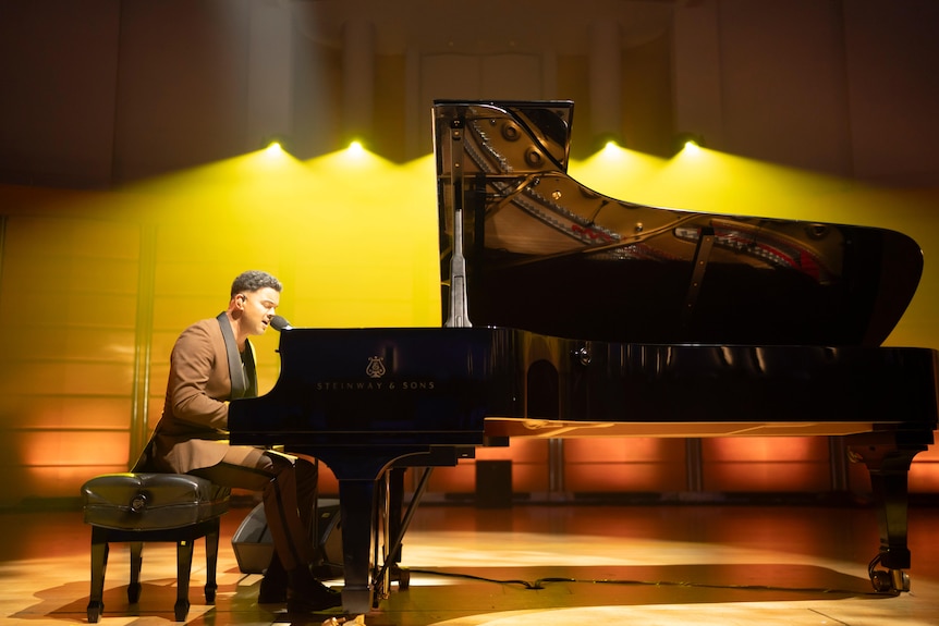 Guy Sebastian sits behind a black grand piano on a stage lit with orange light, singing into a microphone