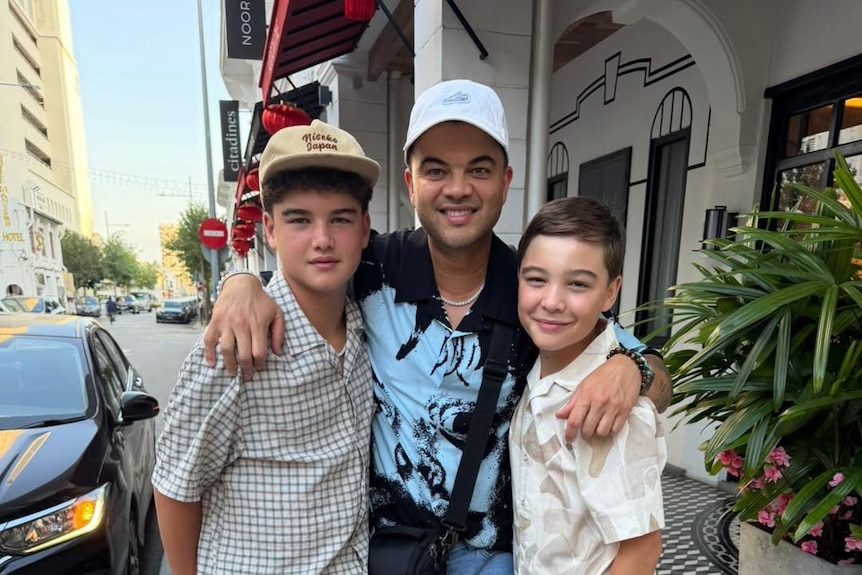 Guy Sebastian stands on a street wearing a cap and smiling, his arms around two young boys