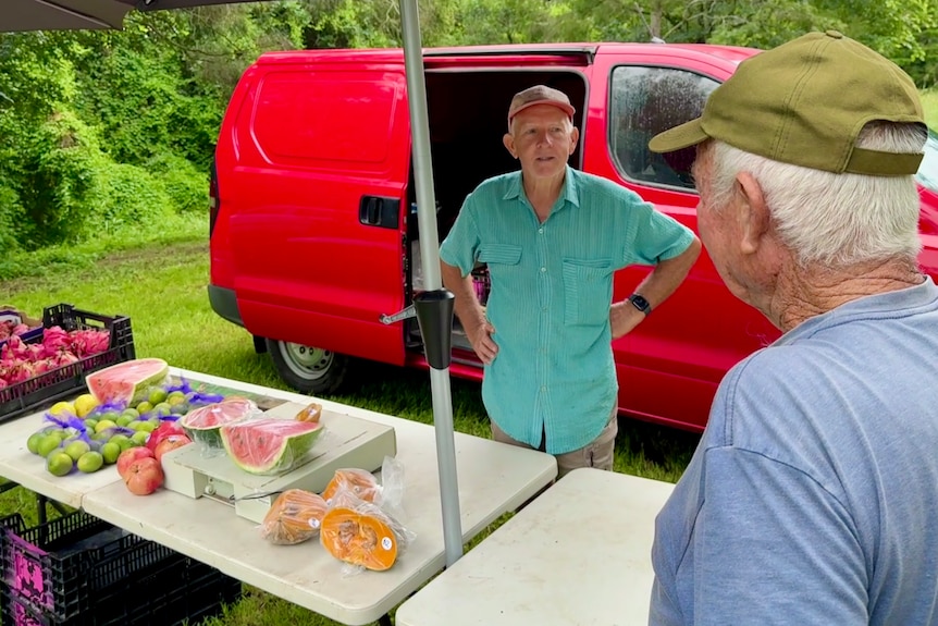 A man stands behind his market stall talking to a customer.