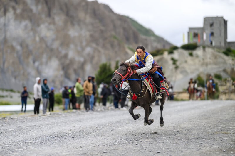 A man in traditional clothing rides a galloping horse along a gravel road, with blurred spectators and rocky mountains in the background.