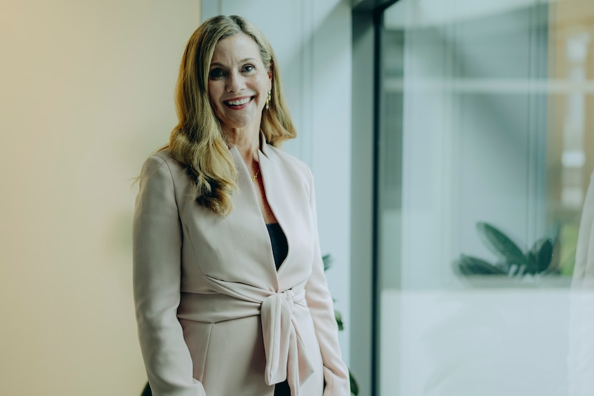 A smiling blonde woman in a light-coloured coat stands near a large window in an office building.