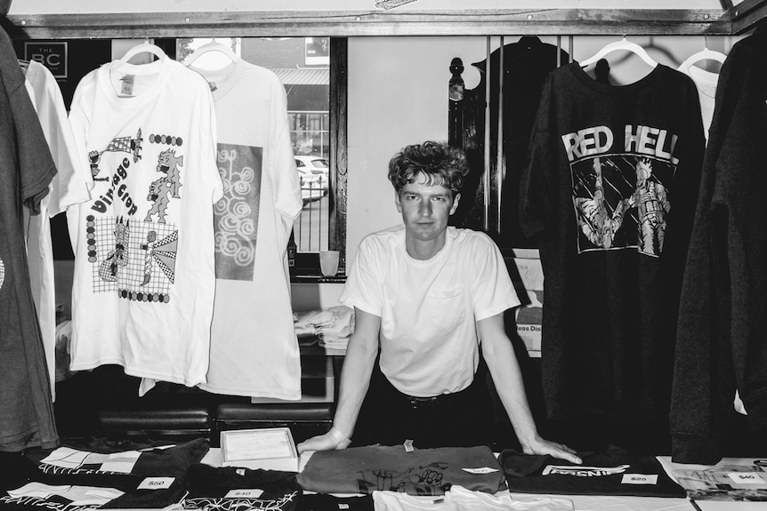 Black and white photo of man stands smiling with hands on table of music and band merchandise inside a pub