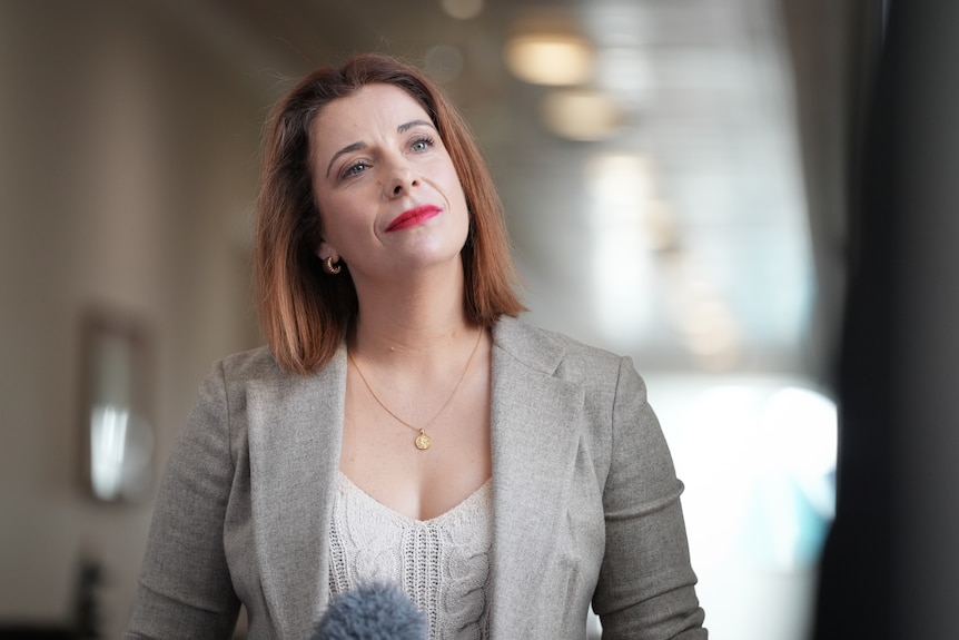 A red-haired woman in a light-coloured blazer stands in a corridor.