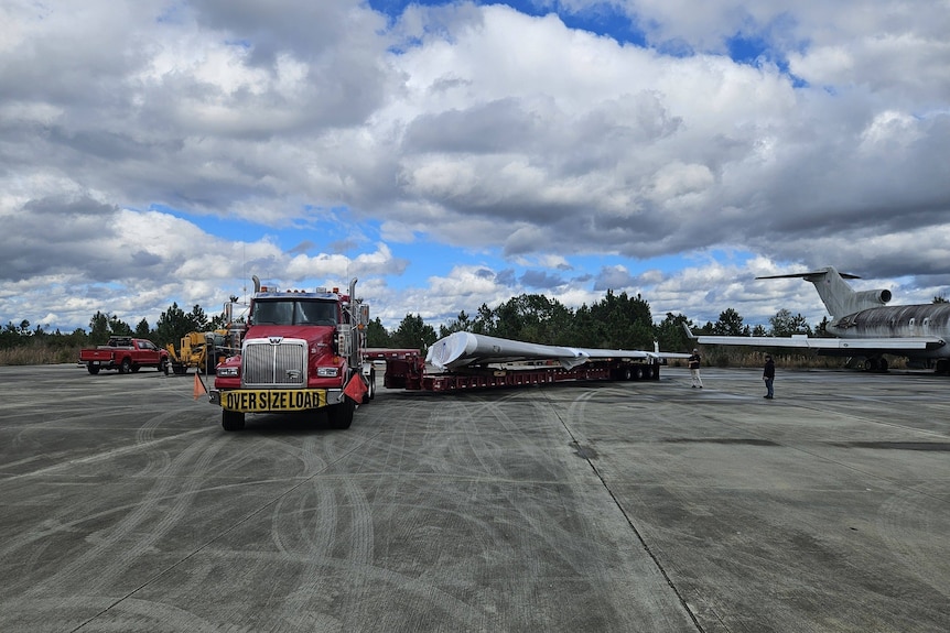 A red truck with a long trailer sits parked with an aircraft wing on the trailer.