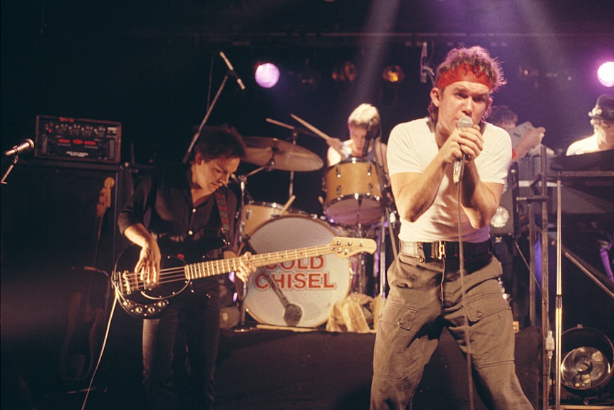 Cold Chisel band performs, circa 1980s. Singer Jimmy Barnes right of frame, in focus, holds mic while wearing red bandana.