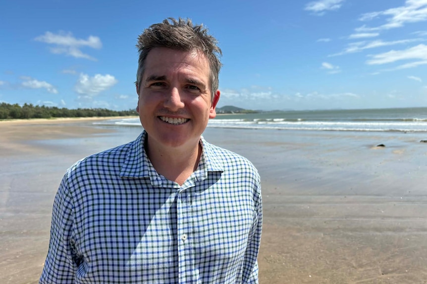A smiling man with short, grey hair wears a checkered shirt while standing on a beach.