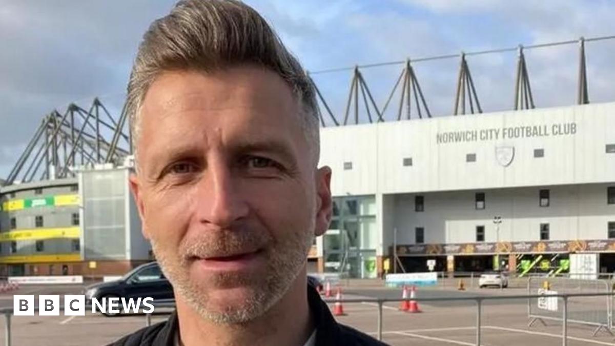 A head-and-shoulders image of a man with grey stubble and grey hair in a quiff. He is standing outside, with Norwich City's Carrow Road stadium behind him