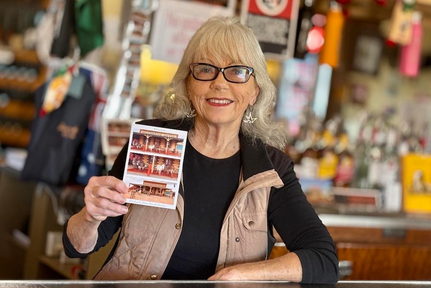 A woman with white hair stands behind a pub counter holding a postcard with donkeys on it.
