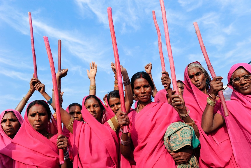 A group of Indian women wearing bright pink saris, holding pink bamboo sticks