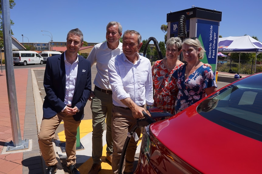 5 people smiling to the left standing outside, Roger Cook holds a EV charger to an electric car 