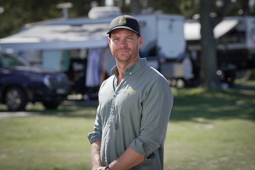 A man with a button up shirt and cap in a caravan park.
