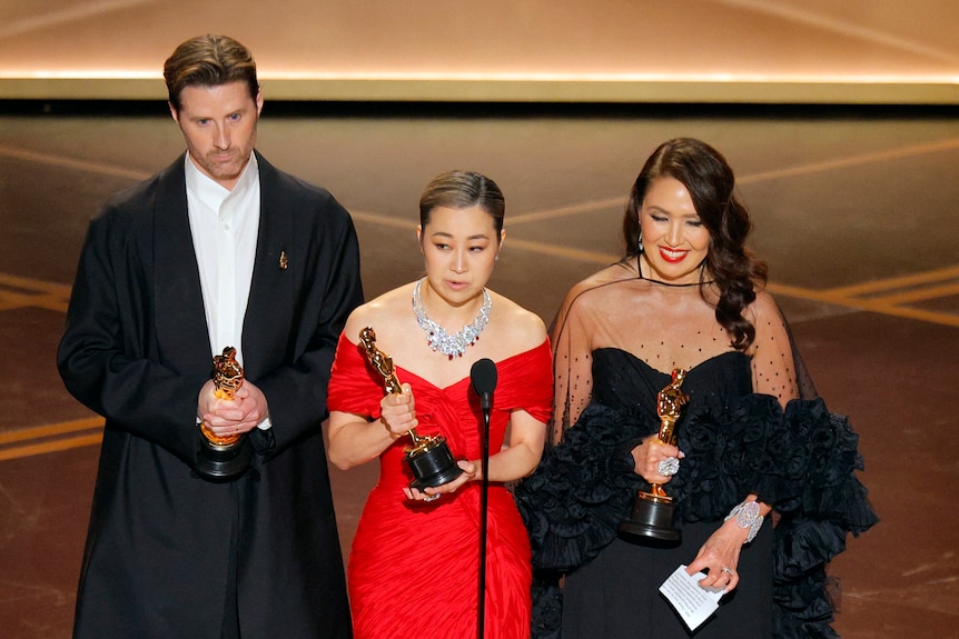 A man and two women in formal clothing stand behind a microphone holding Oscars statuettes.