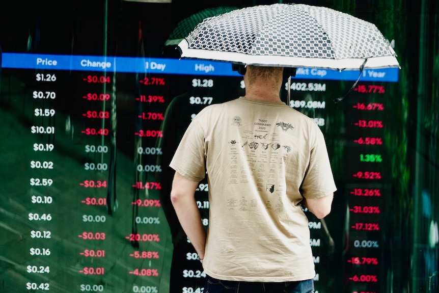 A man stands under an umbrella and looks at a screen showing a financial market falling.