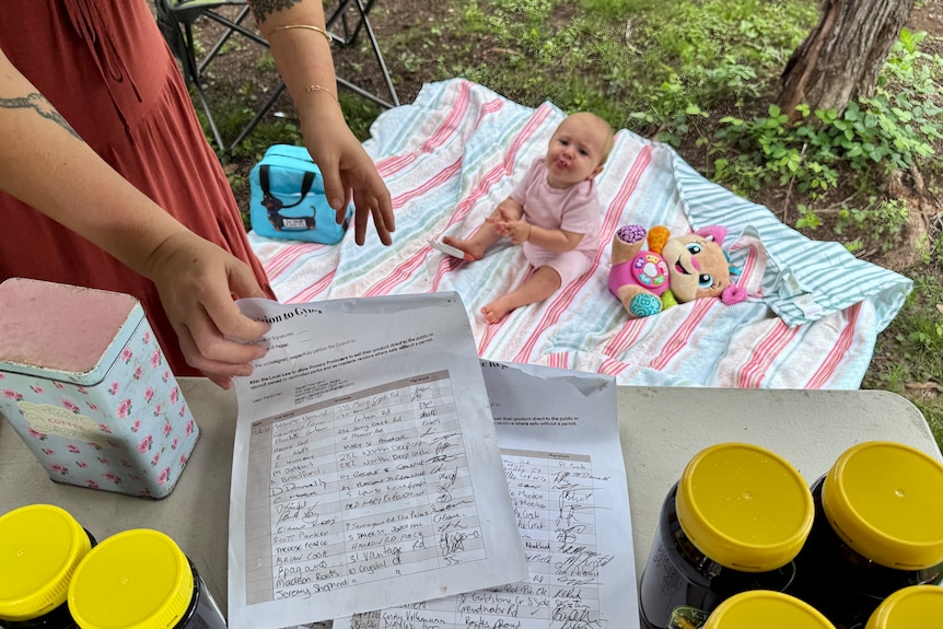 A baby sits next to a rug as hands show a petition on a table above her.