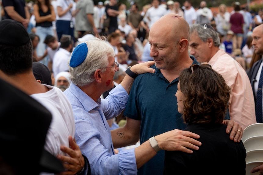 Two men and a woman embrace, a crowd of people behind them. One man is wearing a kippah; the other has a scar on his head.