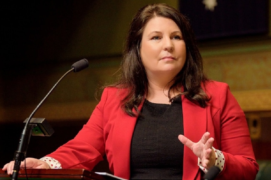 A woman stands in front of a microphone with the Australian Flag hanging on a wall behind her.