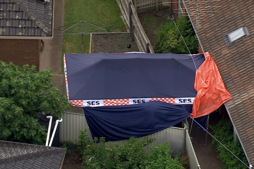 An orange tarp hangs between a brown tiled roof and a blue tent that has "SES" written on it and extends over a fence.