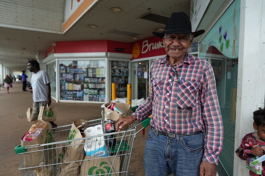 Man with black hat stands next to full trolley outside shops
