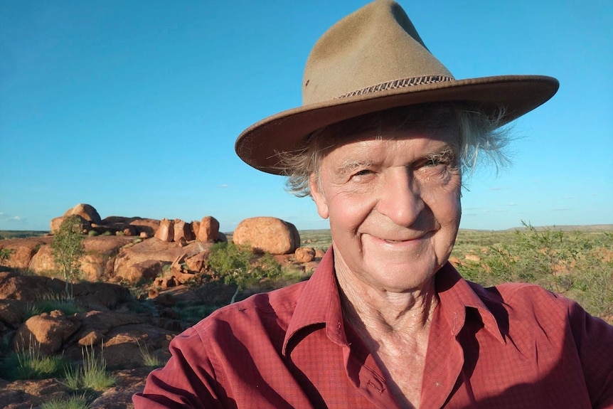 A man in his 70s with a broad hat stands in front of some boulders in the outback, with blue sky overhead.