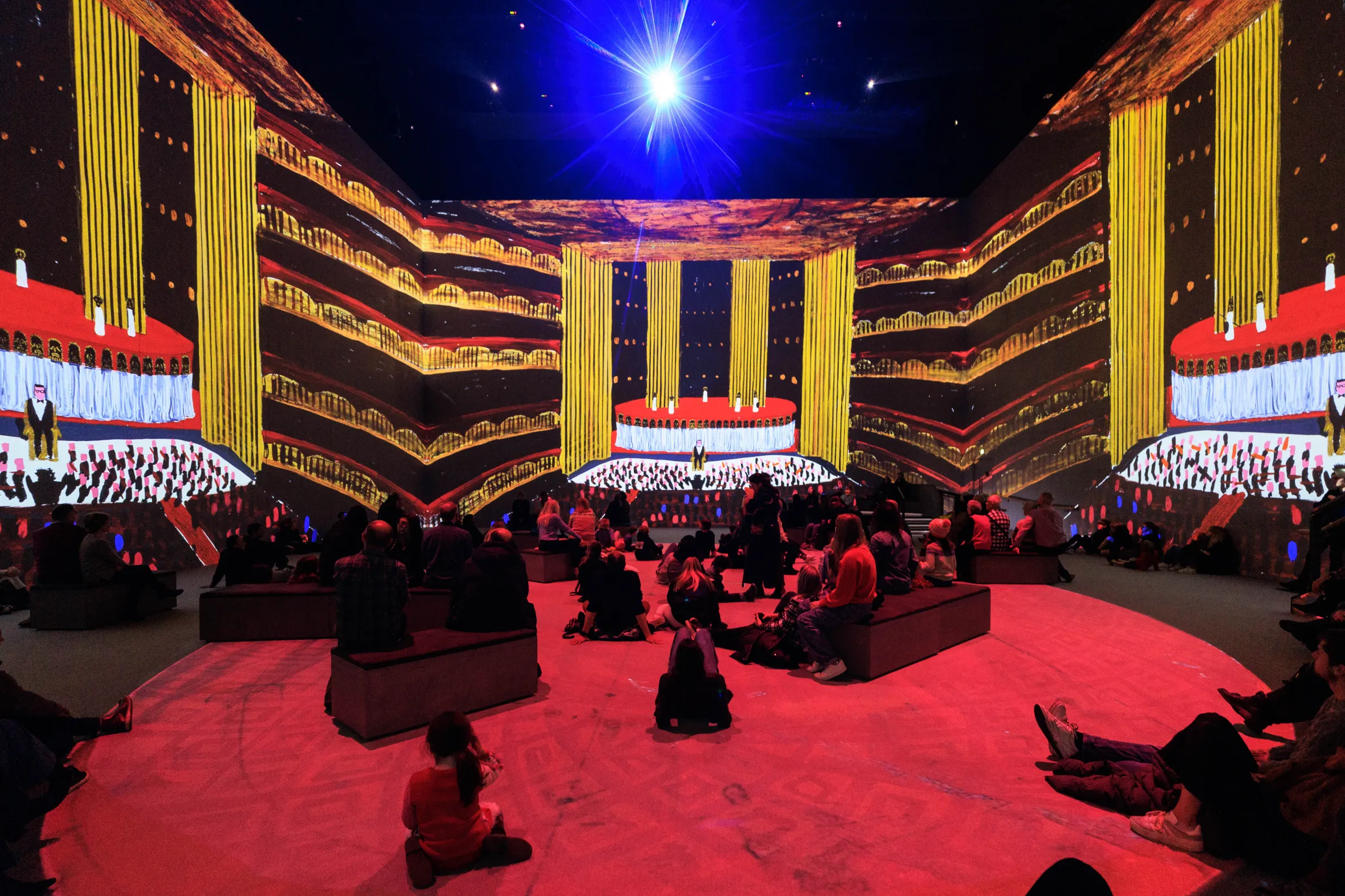 An audience views a projected image of a stage with masks, narrator, and auditorium from David Hockney's "Oedipus Rex" at Lightroom King's Cross.