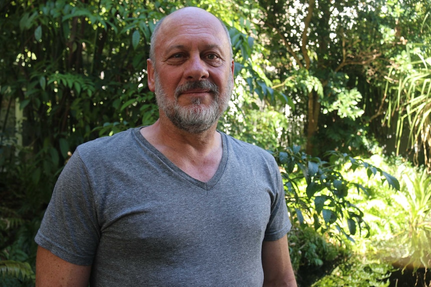 A middle-aged man with a light grey beard and balding head in a t-shirt standing in front of green leafy garden.