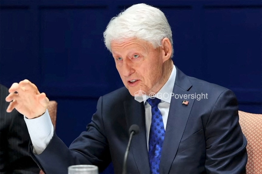Bill Clinton dressed in a suit sits in a seat speaking in front of a microphone.