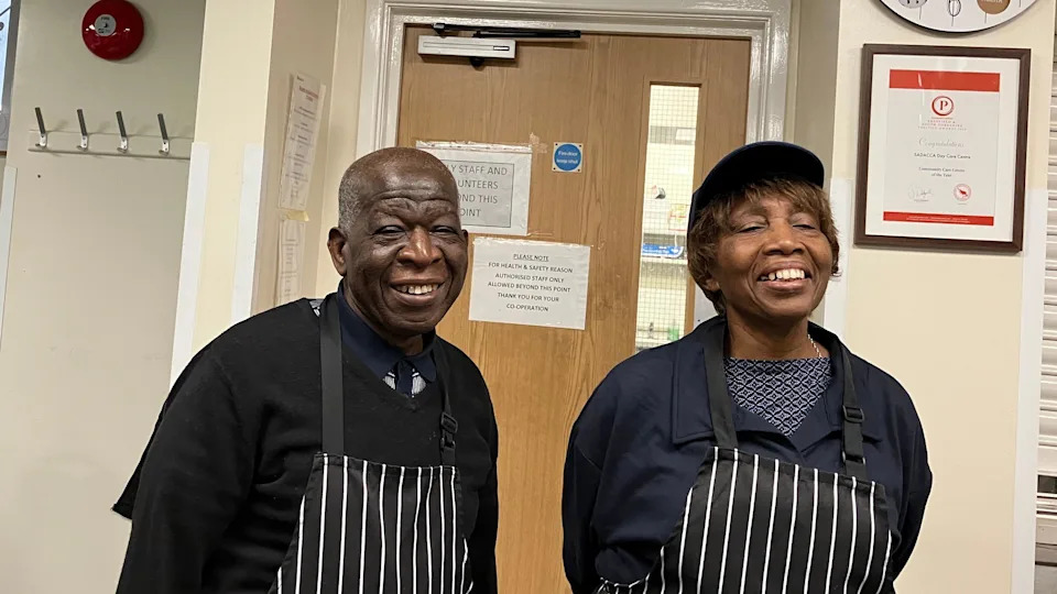 Two older smiling charity volunteers, Webster Modest on the left has short cut grey hair and wears a dark blue polo shirt under a black v-neck jumper, and a black white striped apron.  Pauline Simmonds on the right is smiling broadly has short brown hair visible under and is wearing a blue baseball cap.  Pauline too wears a black and white striped apron, over a blue and white pattern top, and a navy blue shirt style top. 