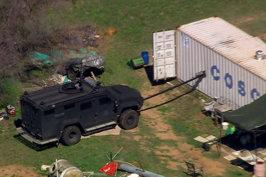 A black police armourned vehicle is parked next to a shipping container. A black pipe is connected between the two.
